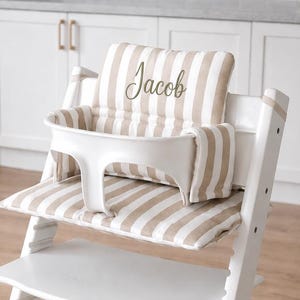 May include: A white wooden high chair featuring a beige and white striped cushion set with a backrest and seat pad. The backrest cushion is embroidered with the name Jacob in a cursive script. The chair includes a white plastic safety guard.
