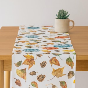 Peut inclure: Chemin de table blanc avec un motif aquarelle de feuilles d'automne, de citrouilles, de glands et de pommes de pin. Le chemin de table est posé sur une table en bois, avec une petite plante succulente en pot en arrière-plan.