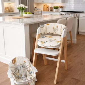 May include: A wooden high chair with a white seat and backrest, featuring a cream-colored cushion adorned with a whimsical chicken pattern. A matching booster seat is on the floor. The chair has a natural wood frame and is set in a bright kitchen.