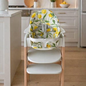 May include: A white high chair with a wooden frame and two white footrests. The seat and backrest feature a lemon and leaf pattern on a white background. The high chair has a safety harness.
