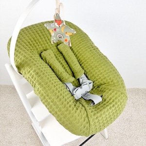 May include: A green waffle-textured baby seat with a white wooden frame. A star-shaped toy with a fox and rainbow pattern hangs above the seat. The seat has gray straps for safety.