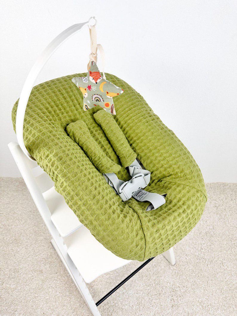 May include: A green waffle-textured baby seat with a white wooden frame. A star-shaped toy with a fox and rainbow pattern hangs above the seat. The seat has gray straps for safety.