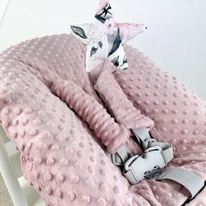 May include: A pink baby seat cushion with a textured surface and a matching headrest. A star-shaped toy with a floral pattern hangs above the seat. The seat has a gray safety harness.