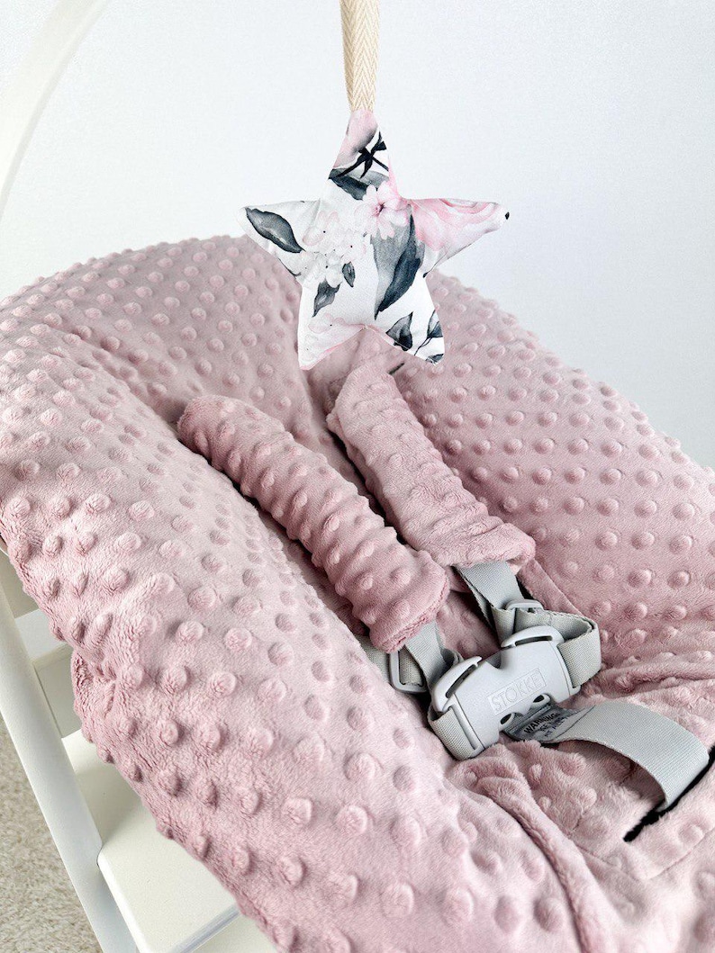 May include: A pink baby seat cushion with a textured surface and a matching headrest. A star-shaped toy with a floral pattern hangs above the seat. The seat has a gray safety harness.