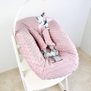 May include: A pink baby seat cover with a textured, dotted surface, featuring a gray safety harness. A white arch supports a star-shaped mobile with a floral pattern. The seat is on a white wooden high chair.