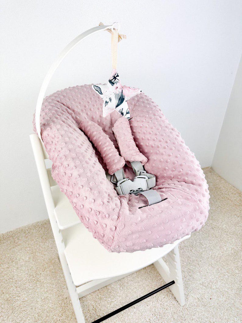 May include: A pink baby seat cover with a textured, dotted surface, featuring a gray safety harness. A white arch supports a star-shaped mobile with a floral pattern. The seat is on a white wooden high chair.