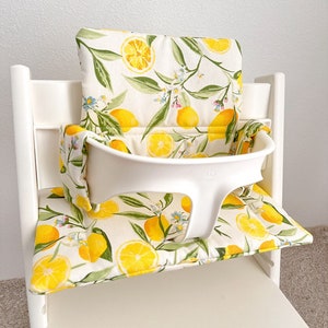 Wipeable high chair cushion compatible with Stokke Tripp Trapp, shown on wooden chair with modern dining table, soft padded seat cover in neutral color.