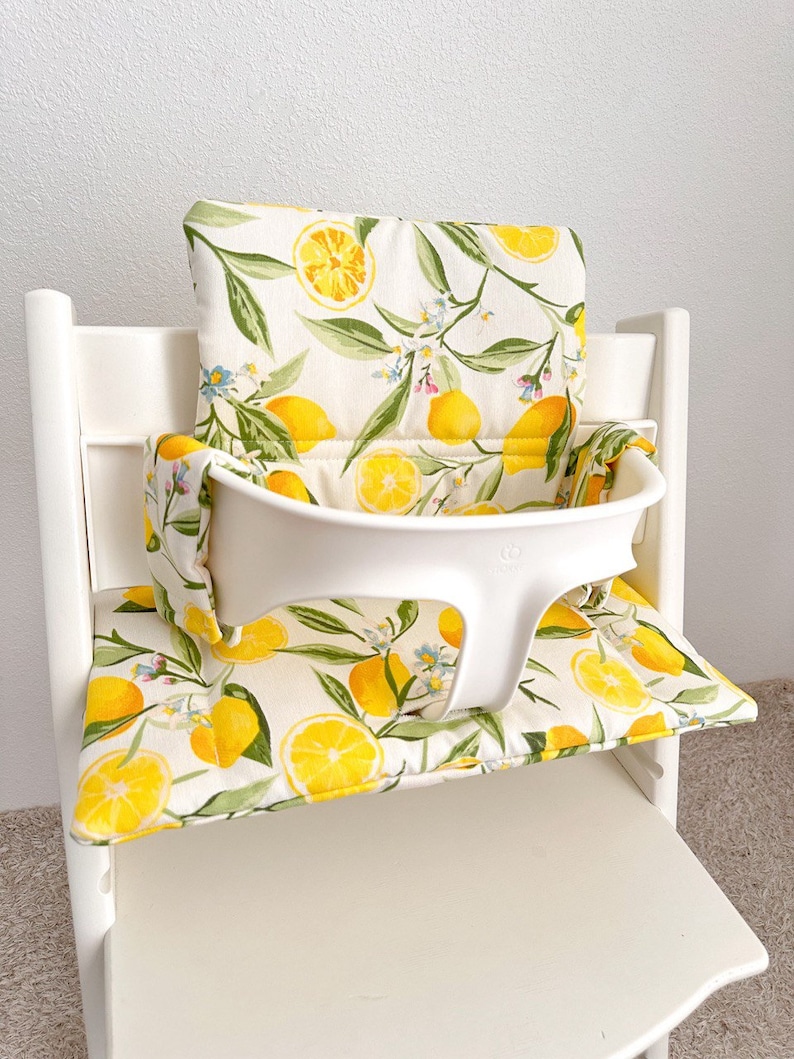 Wipeable high chair cushion compatible with Stokke Tripp Trapp, shown on wooden chair with modern dining table, soft padded seat cover in neutral color.