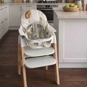 May include: A modern high chair with a white frame and natural wood legs. The seat and backrest feature a cream-colored fabric with a repeating chicken pattern. The chair has a tray and safety straps for a baby or toddler.