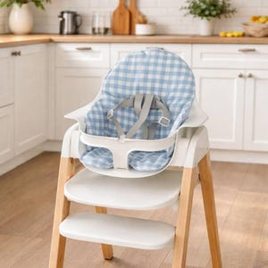 May include: A white and wood-toned high chair with a blue and white gingham patterned seat. The high chair has a safety harness and a tray. The legs are made of light-colored wood.