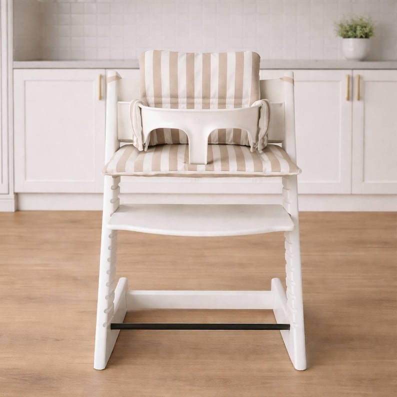 May include: A white wooden high chair with a beige and white striped cushion and backrest. The chair has a white tray and a black footrest. The high chair is designed for infants and toddlers.