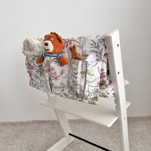 Puede incluir: Silla alta blanca con un organizador de tela con estampado floral sujeto a la parte posterior. El organizador tiene varios bolsillos y est&aacute; lleno de animales de peluche y una toallita para beb&eacute;s.