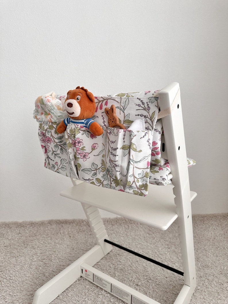 Puede incluir: Silla alta blanca con un organizador de tela con estampado floral sujeto a la parte posterior. El organizador tiene varios bolsillos y est&aacute; lleno de animales de peluche y una toallita para beb&eacute;s.