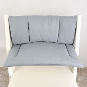 May include: A white high chair with a light gray padded seat and back. The chair has a curved back with three separate cushions.