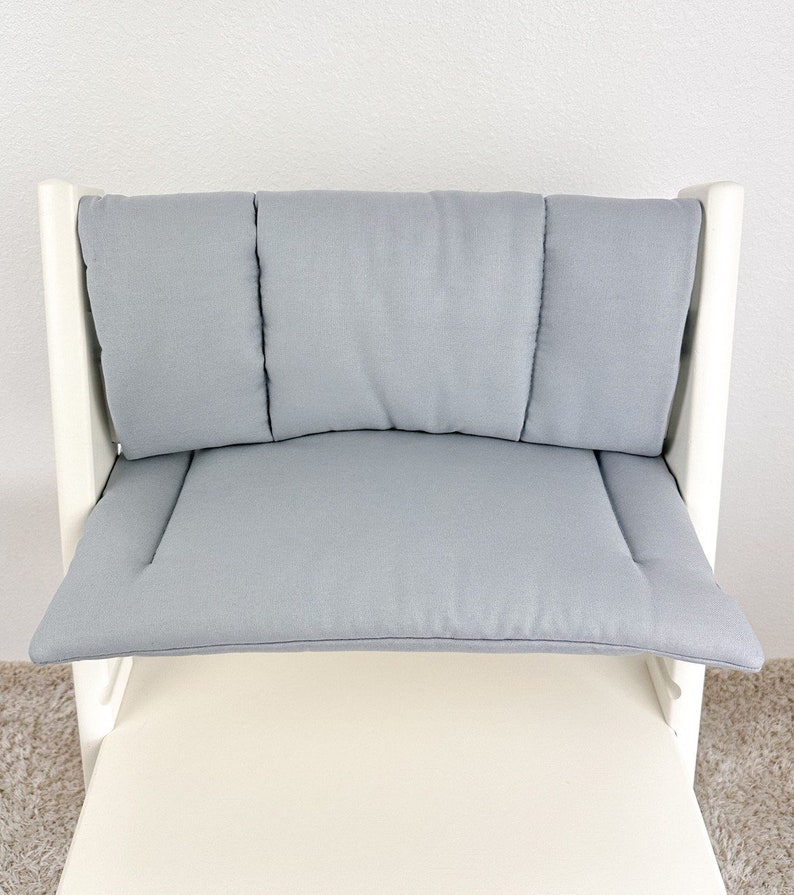 May include: A white high chair with a light gray padded seat and back. The chair has a curved back with three separate cushions.