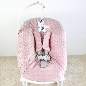 May include: A pink baby seat cover with a textured, dotted pattern. The seat has a gray safety harness and is adorned with a hanging star-shaped mobile. The star is patterned with floral and watercolor designs. The seat is on a white wooden chair.