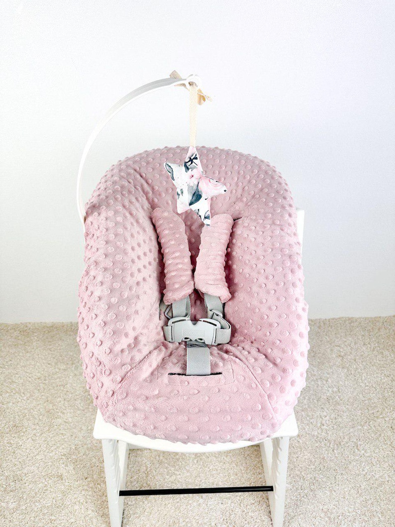 May include: A pink baby seat cover with a textured, dotted pattern. The seat has a gray safety harness and is adorned with a hanging star-shaped mobile. The star is patterned with floral and watercolor designs. The seat is on a white wooden chair.