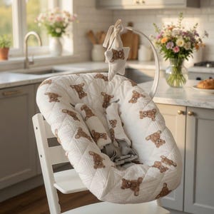 May include: A white high chair with a quilted seat cover featuring a repeating pattern of brown teddy bears. The chair has a white frame and a gray safety harness. A star-shaped toy hangs above the seat.