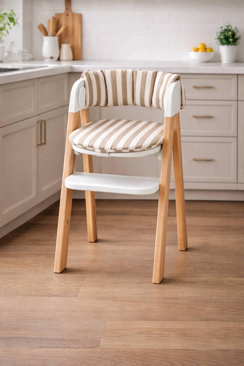 May include: A modern high chair with a light wood frame and white accents. The seat and backrest feature a beige and white striped pattern. The chair has a footrest and is designed for young children.