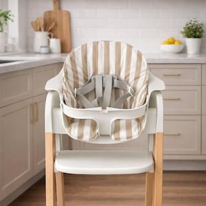 May include: A white high chair with a beige and white striped seat cushion and a gray safety harness. The chair has light wood legs and is designed for a baby or toddler. The high chair is in a kitchen setting.