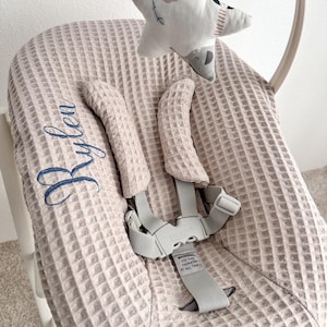 Könnte beinhalten: Ein hellbeige Babywippe mit einem strukturierten Waffelstoff. Die Wippe hat einen grauen Sicherheitsgurt und ein hängendes sternförmiges Mobile. Der Name "Rylen" ist in Blau auf den Stoff gestickt.