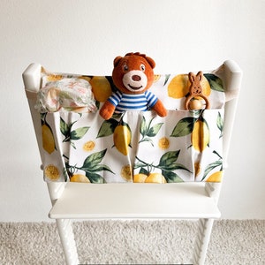 May include: White high chair with a lemon-patterned fabric organizer attached. The organizer has three pockets, one with a stuffed bear, one with a stuffed bunny, and one with a cloth diaper.