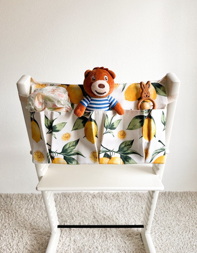 May include: White high chair with a lemon-patterned fabric organizer attached. The organizer has three pockets, one with a stuffed bear, one with a stuffed bunny, and one with a cloth diaper.