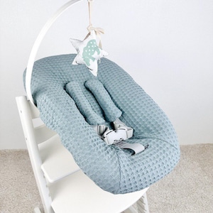 May include: A baby high chair with a light blue waffle-textured seat cover and a white frame. A white arch holds a hanging star-shaped toy with a light green pattern. The chair has a safety harness.
