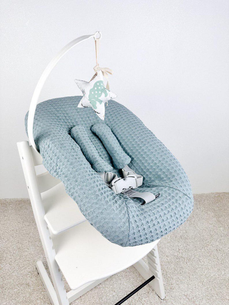 May include: A baby high chair with a light blue waffle-textured seat cover and a white frame. A white arch holds a hanging star-shaped toy with a light green pattern. The chair has a safety harness.