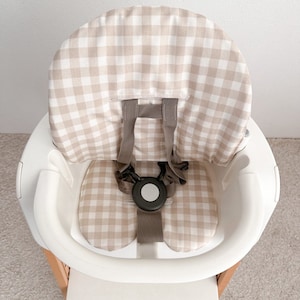 K&ouml;nnte beinhalten: Ein wei&szlig;er Hochstuhl mit einer beige-wei&szlig;en Gingham-Muster-Bezug. Der Stuhl hat einen F&uuml;nf-Punkt-Gurt und ein Tablett.