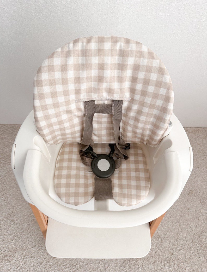 K&ouml;nnte beinhalten: Ein wei&szlig;er Hochstuhl mit einer beige-wei&szlig;en Gingham-Muster-Bezug. Der Stuhl hat einen F&uuml;nf-Punkt-Gurt und ein Tablett.