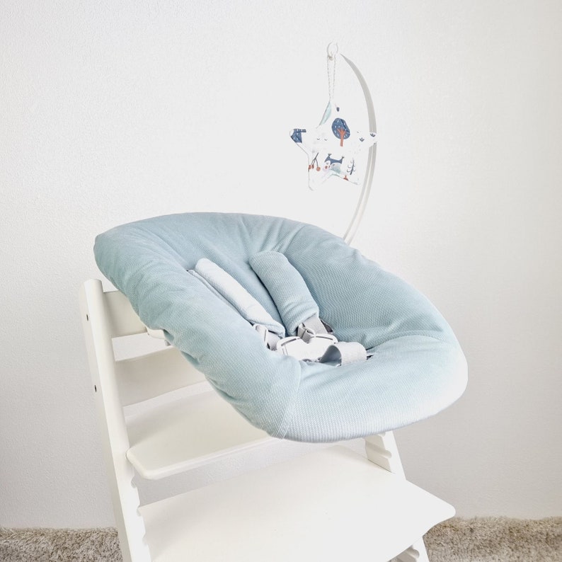 May include: A light blue baby seat cushion with a safety harness, placed on a white wooden high chair. A decorative mobile with a star-shaped design and woodland illustrations hangs above the seat.