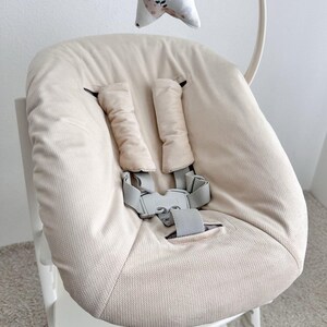 May include: Beige high chair seat with a five-point harness and padded shoulder straps. The seat has a soft, textured fabric and a gray buckle. A decorative star-shaped toy hangs above the seat.