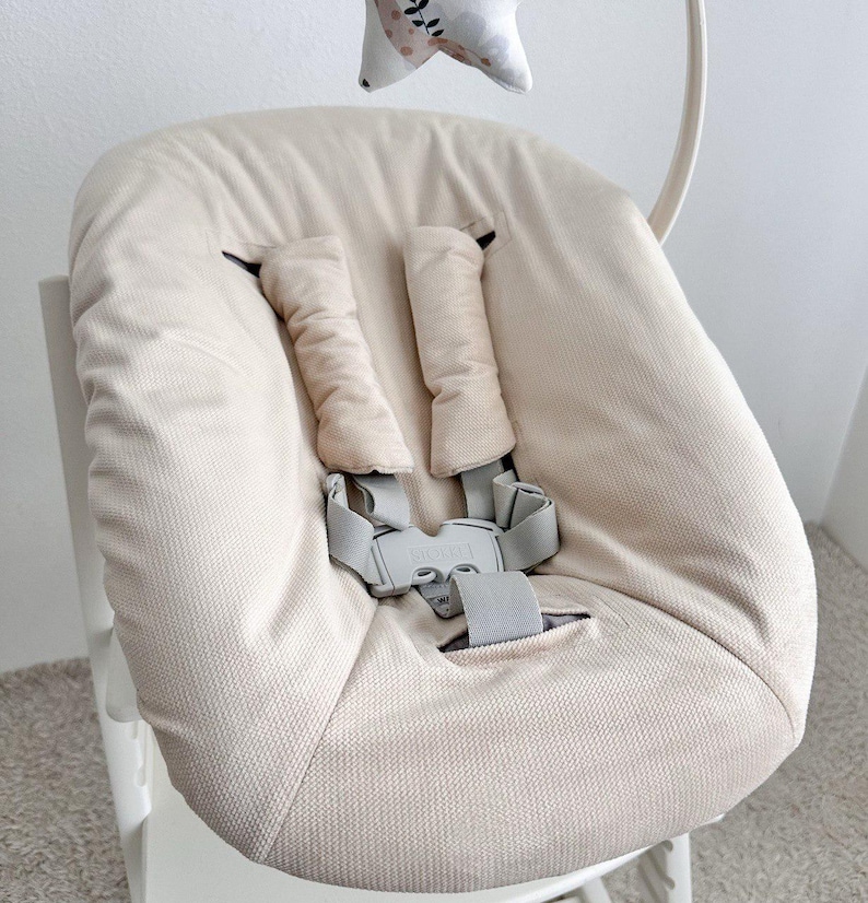 May include: Beige high chair seat with a five-point harness and padded shoulder straps. The seat has a soft, textured fabric and a gray buckle. A decorative star-shaped toy hangs above the seat.