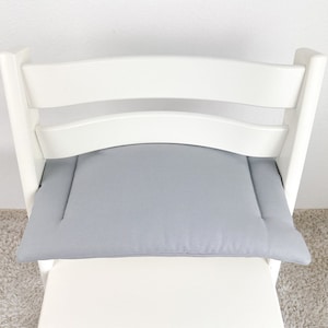 May include: A gray padded seat cushion for a high chair. The cushion is made of a soft, textured fabric and has a simple, rectangular shape.