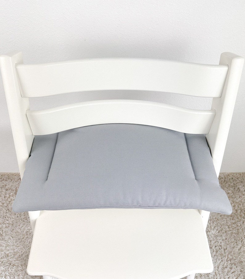 May include: A gray padded seat cushion for a high chair. The cushion is made of a soft, textured fabric and has a simple, rectangular shape.