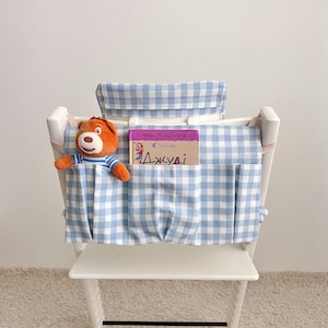 May include: A white high chair with a blue and white gingham fabric organizer. A stuffed bear wearing a striped shirt and a book with text in a foreign language are in the organizer's pockets.