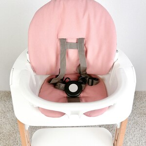 May include: A pink high chair with a white tray and a wooden base. The chair has a pink padded seat and back, and a five-point harness.