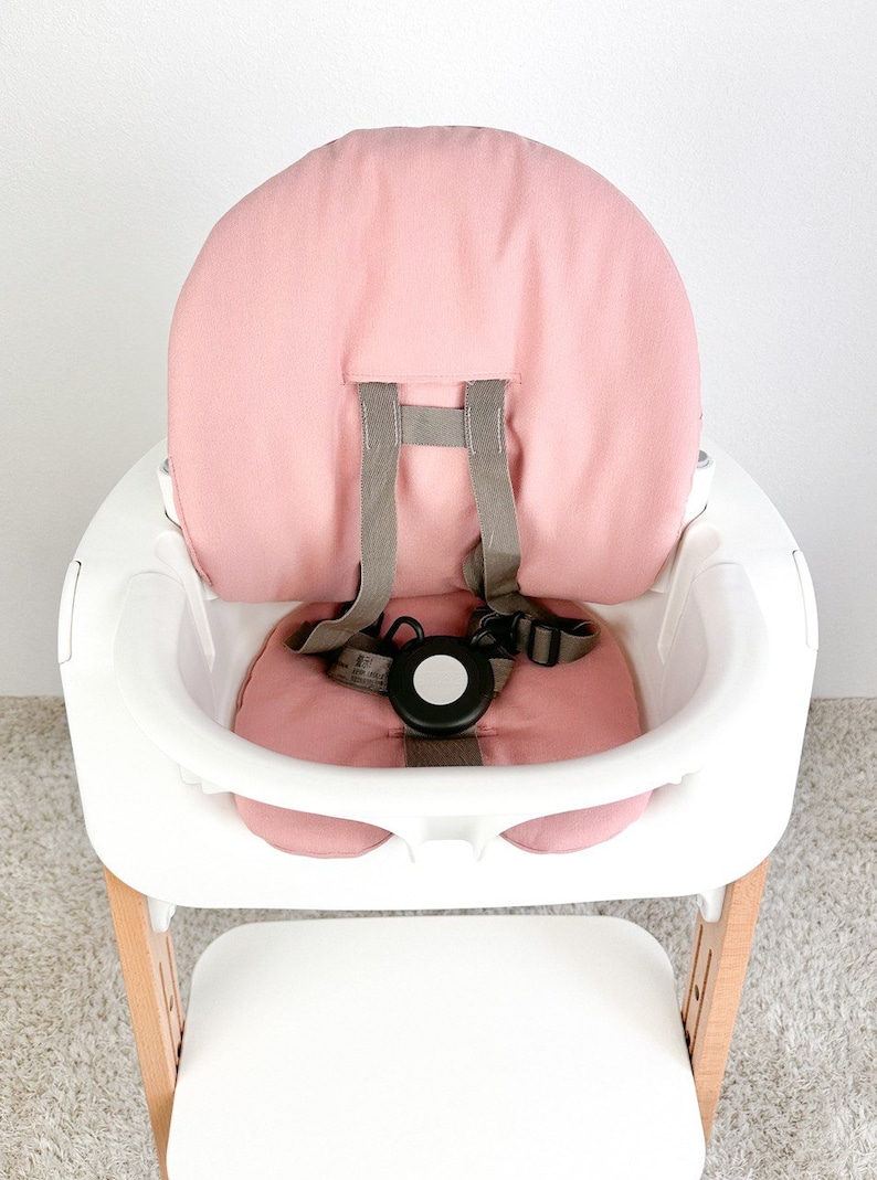 May include: A pink high chair with a white tray and a wooden base. The chair has a pink padded seat and back, and a five-point harness.