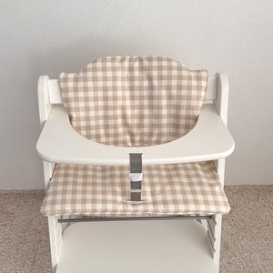 May include: White high chair with a beige and white gingham pattern seat cushion and a gray strap.