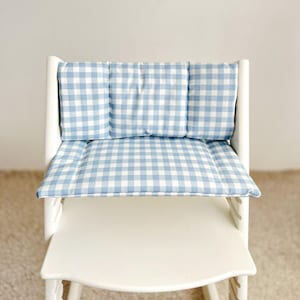 May include: A white high chair with a blue and white gingham cushion set. The cushion set includes a backrest and seat cushion. The fabric has a classic checkered pattern. The chair is designed for a child.