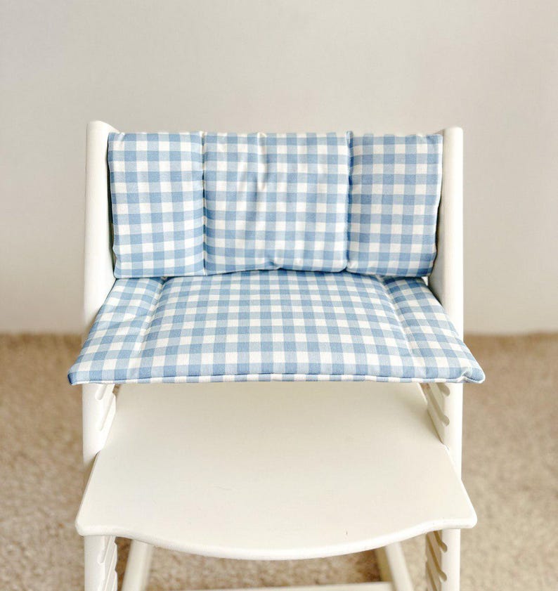 May include: A white high chair with a blue and white gingham cushion set. The cushion set includes a backrest and seat cushion. The fabric has a classic checkered pattern. The chair is designed for a child.
