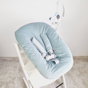 May include: A light blue baby high chair cushion with a safety harness. The cushion is made of a soft, corduroy-like material. A white wooden high chair frame supports the cushion. A decorative mobile with a moon and star design hangs above the chair.