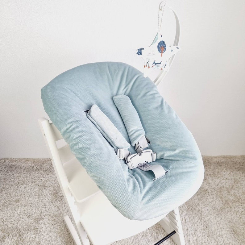 May include: A light blue baby high chair cushion with a safety harness. The cushion is made of a soft, corduroy-like material. A white wooden high chair frame supports the cushion. A decorative mobile with a moon and star design hangs above the chair.