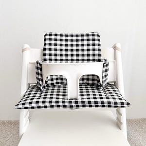 May include: A white high chair with a black and white checkered cushion and backrest. The chair has a white plastic tray and straps. The cushion and backrest are made of a soft fabric.