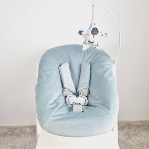 May include: A light blue baby seat with a five-point harness. A white metal arch supports a star-shaped fabric toy with a forest and animal print. The seat is designed for infant comfort and safety.