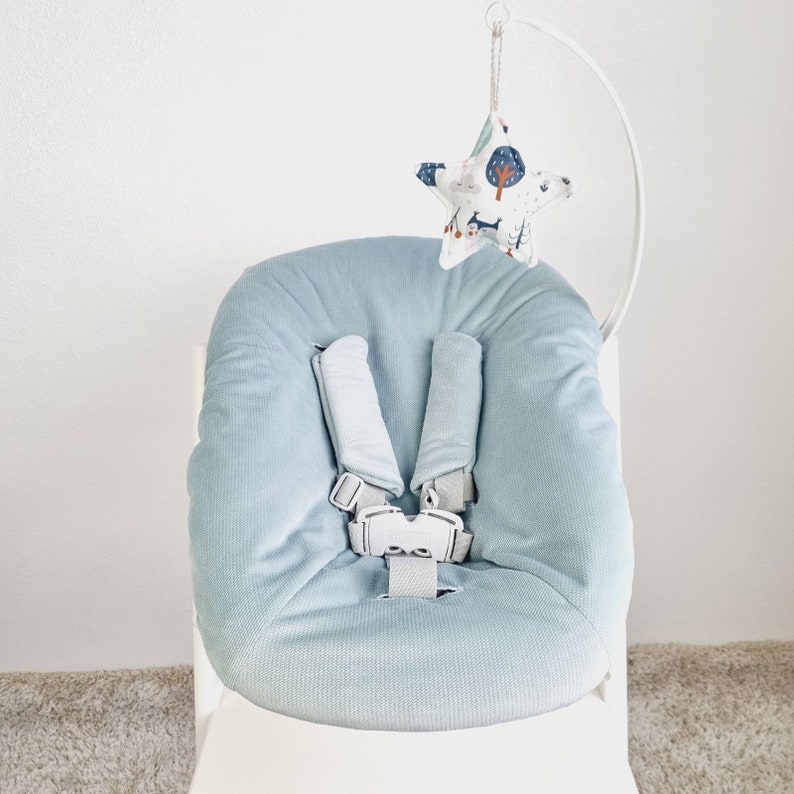 May include: A light blue baby seat with a five-point harness. A white metal arch supports a star-shaped fabric toy with a forest and animal print. The seat is designed for infant comfort and safety.