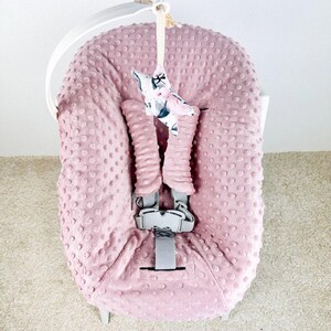 May include: A pink baby car seat cover with a textured, dotted pattern. The cover has a gray harness and a decorative star-shaped toy with a floral print. The car seat is on a white frame.