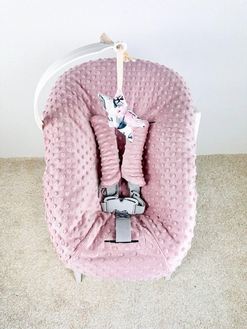 May include: A pink baby car seat cover with a textured, dotted pattern. The cover has a gray harness and a decorative star-shaped toy with a floral print. The car seat is on a white frame.