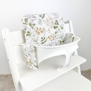 Puede incluir: Silla alta blanca con un coj&iacute;n de asiento estampado floral y una correa a juego. El coj&iacute;n tiene un fondo blanco con hojas verdes, flores amarillas y detalles morados.